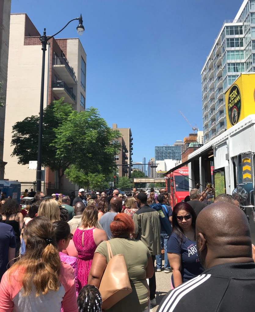 Crowd lined up at food trucks beside city buildings on a clear day