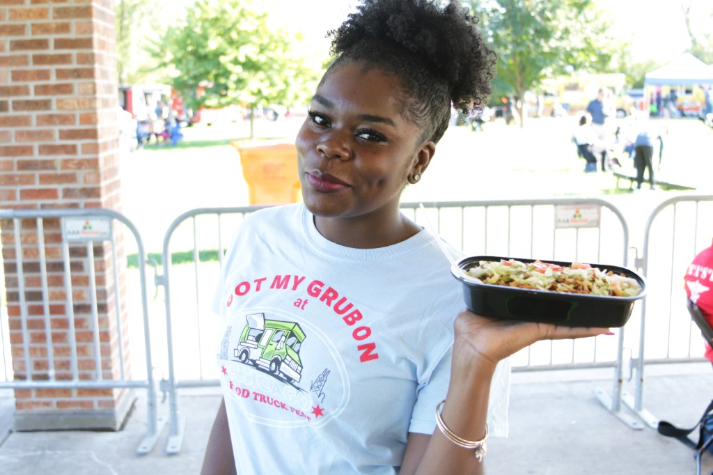 Guest in a Chicago Food Truck Fest shirt holding a takeout bowl at Labagh Woods with festival fencing and trees in the background.