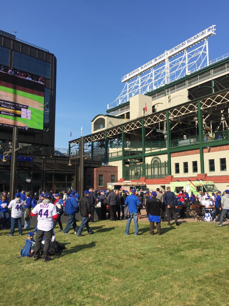 Crowd in Cubs gear on Gallagher Way with food trucks and Wrigley steelwork
