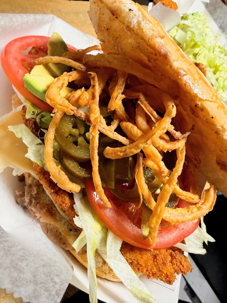 La Calaca — Milanesa torta with haystack onions, crispy breaded cutlet, avocado, tomato, and pickled jalapeños on a toasted roll
